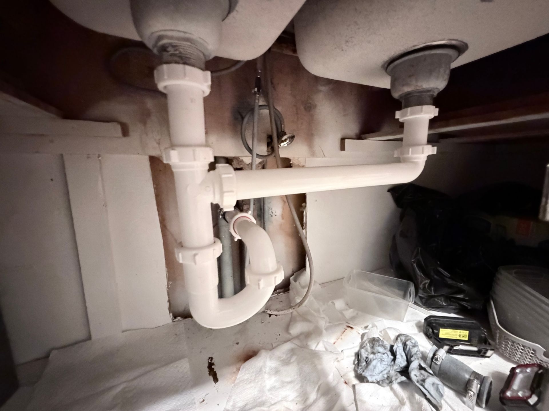 Under-sink plumbing with white pipes and hoses, tools on the cabinet floor, and a dark leak-stained area on the right