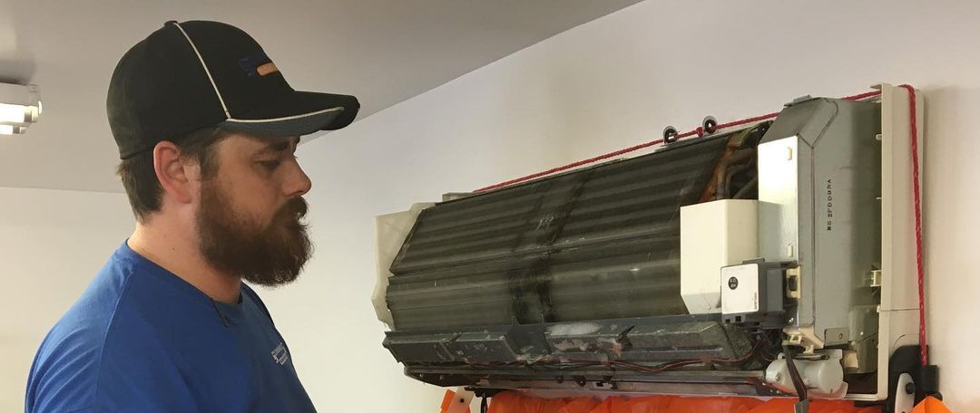 A man with a beard wearing a cap is looking at an air conditioner unit on a wall.