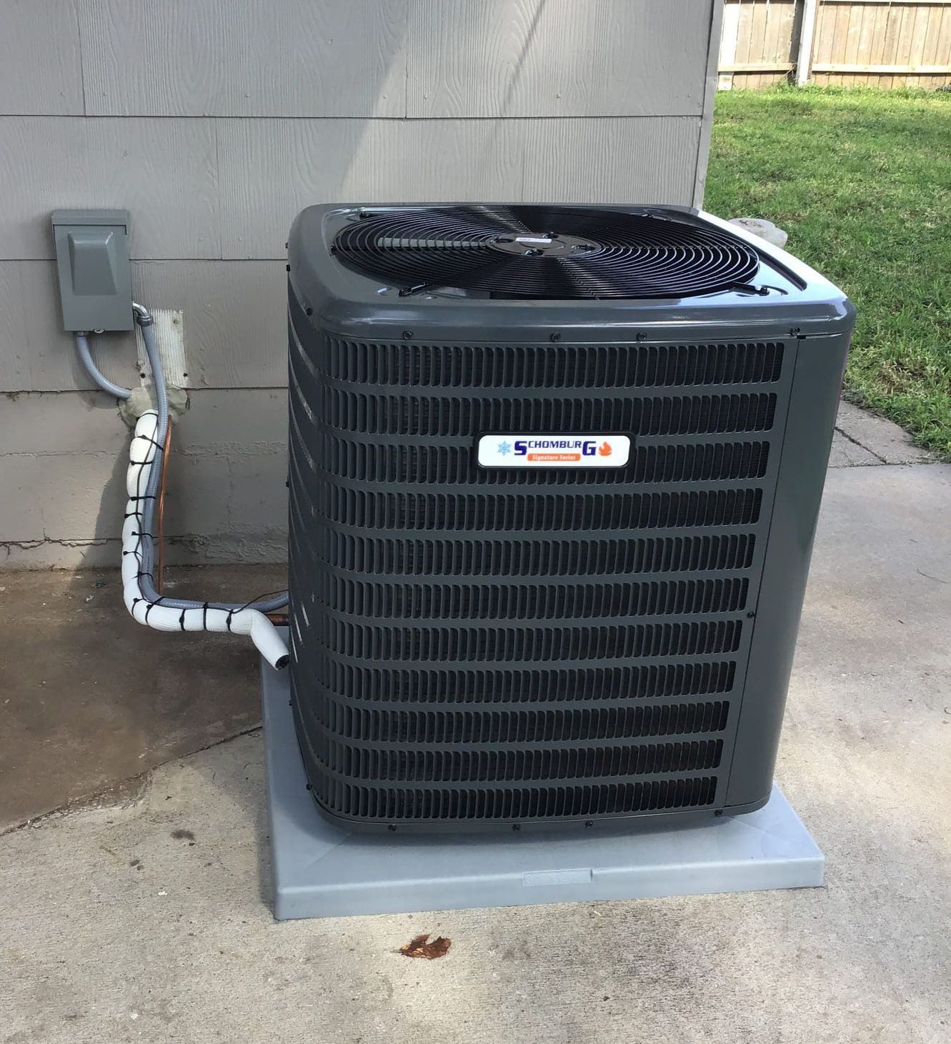 Gray air conditioning unit outside against a gray house wall, on a concrete pad.