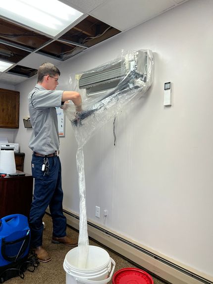 Man installing an air conditioning unit on a wall. The unit is covered in plastic, and there is a bucket below.