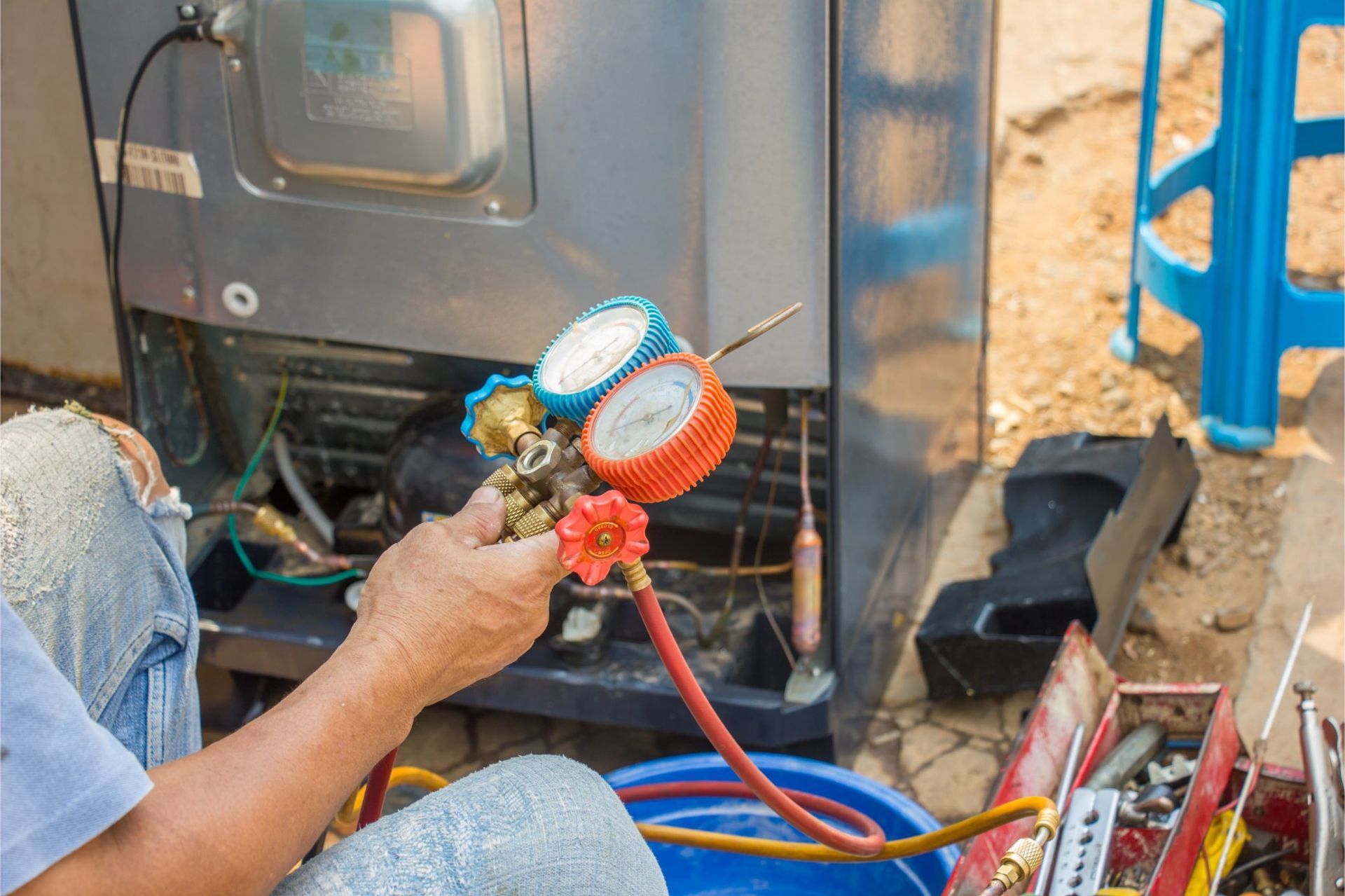 A technician holds a manifold gauge set while repairing the back of a refrigerator outdoors.