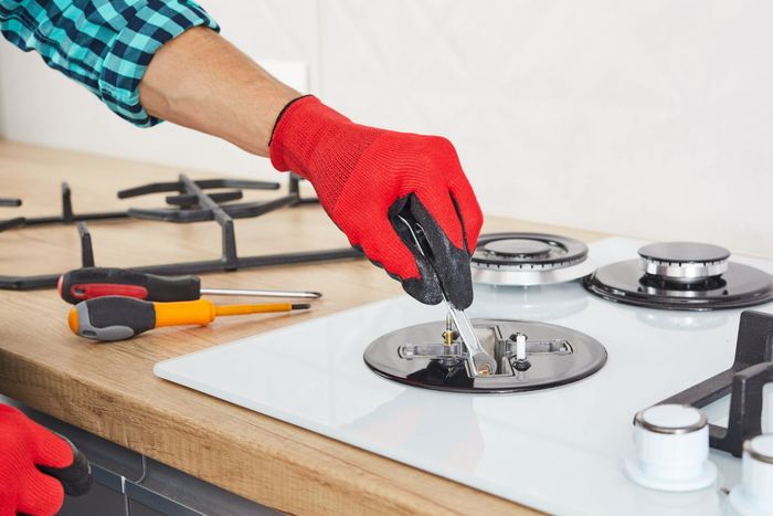 A person wearing red gloves uses a tool to repair a burner on a white glass stovetop in a kitchen.