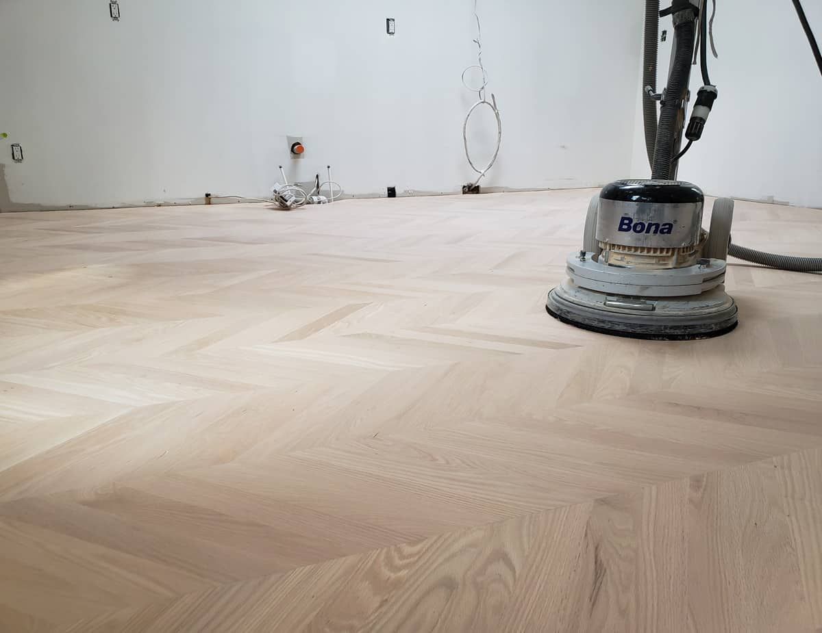 Une ponceuse à plancher sur un plancher en bois clair à motif à chevrons dans une pièce aux murs blancs.