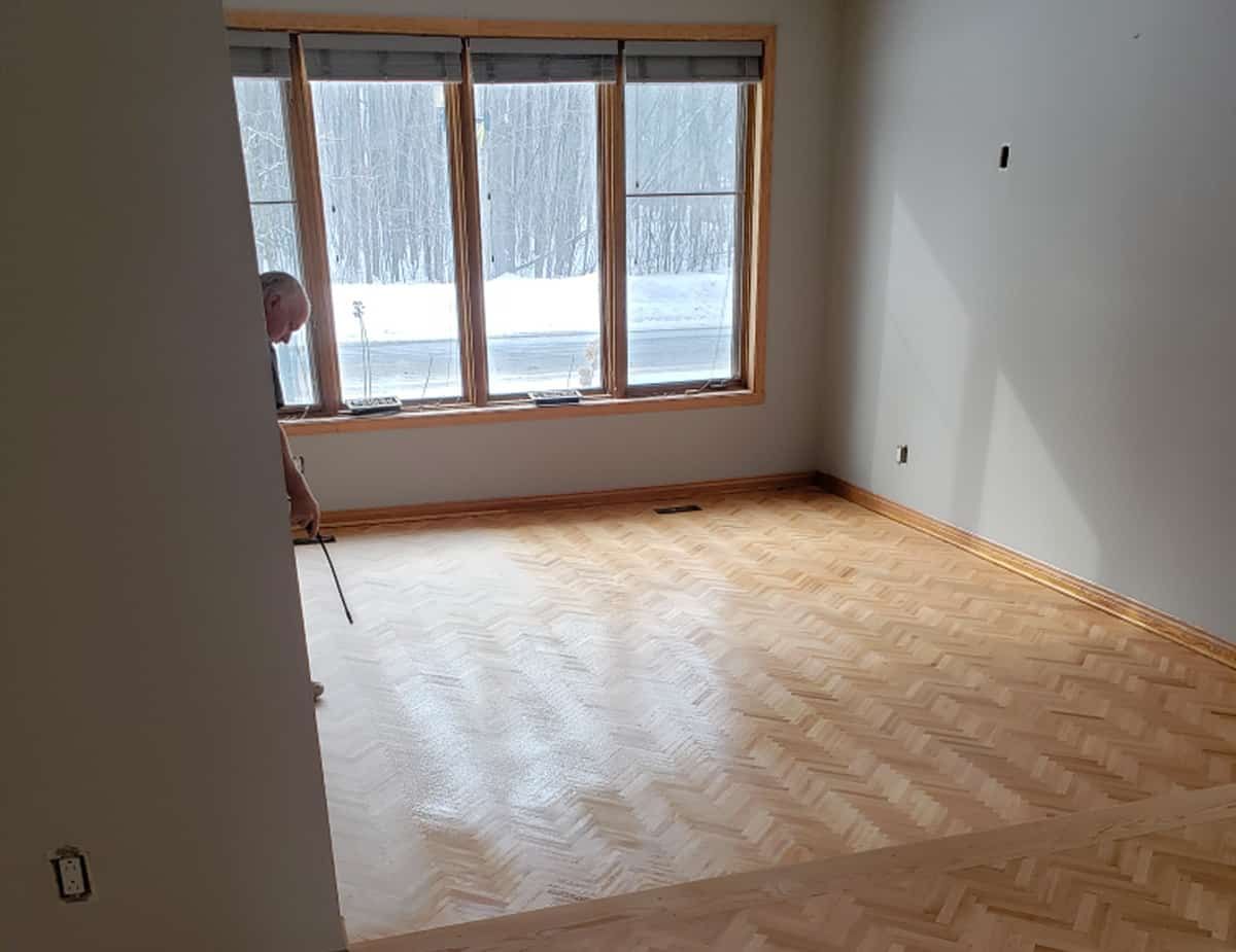 Un homme remet à neuf un parquet en bois clair dans une pièce dont les fenêtres donnent sur un paysage enneigé.