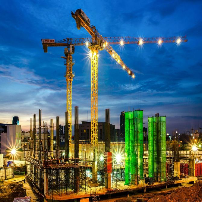 Construction site at dusk with tower cranes, steel frames, and bright green-lit building columns