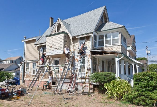 Workers on ladders repairing the exterior of a two-story stone house under a clear blue sky
