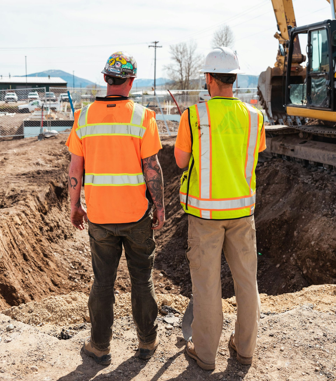 Two workers in safety vests and hard hats standing at a construction site next to heavy machinery.