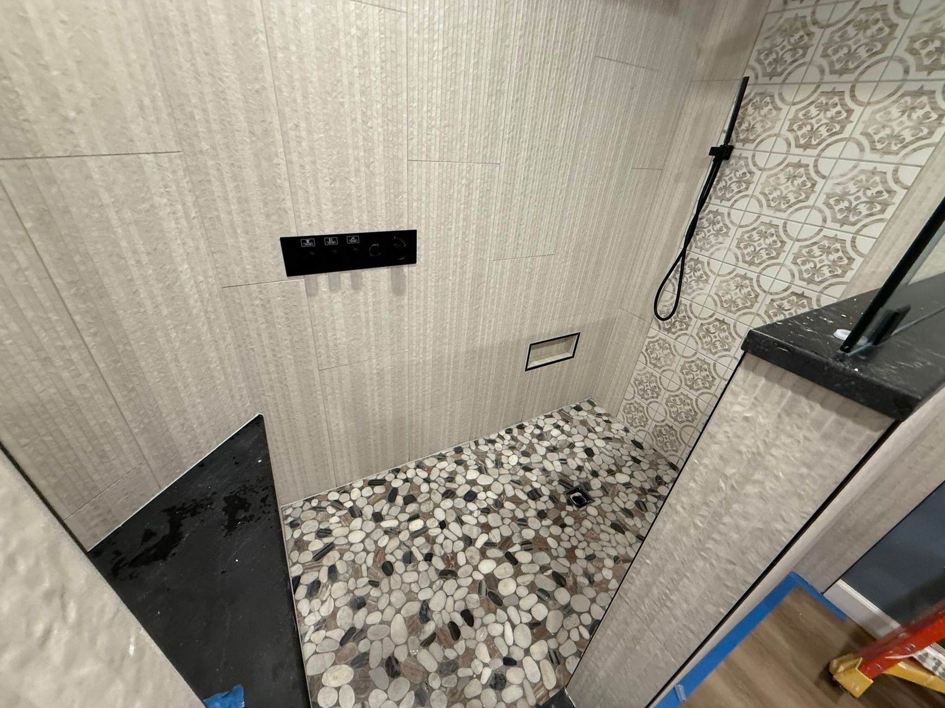 Small tiled shower room with beige walls, pebble floor, wall controls, and a black shower head on the right.