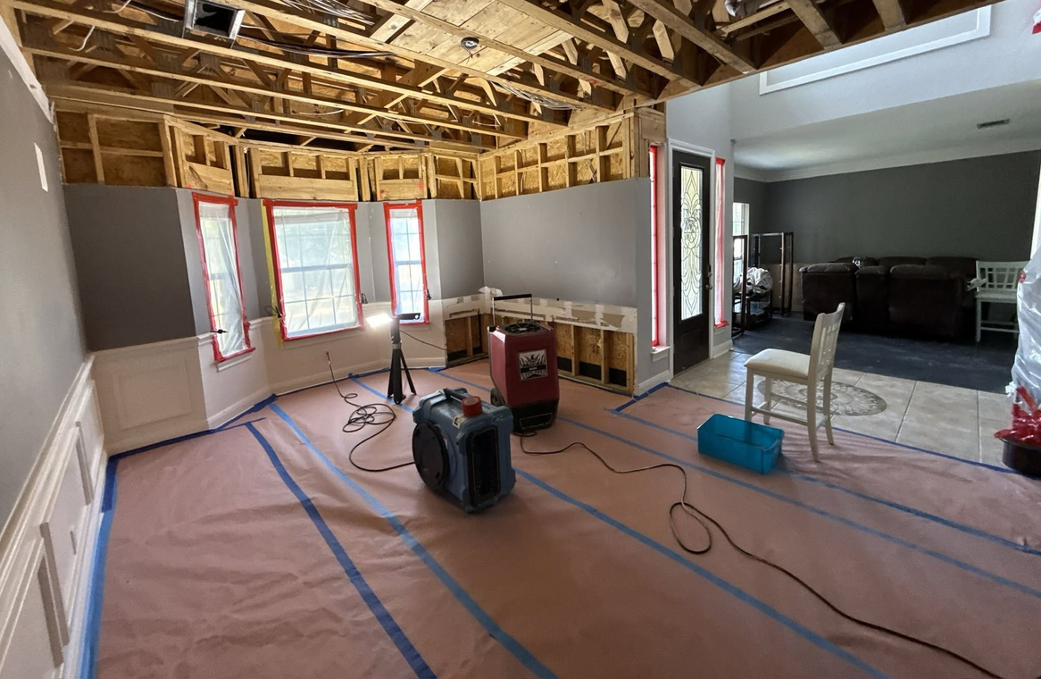 Room under renovation with exposed ceiling beams, covered floor, tools, and a large window bay.