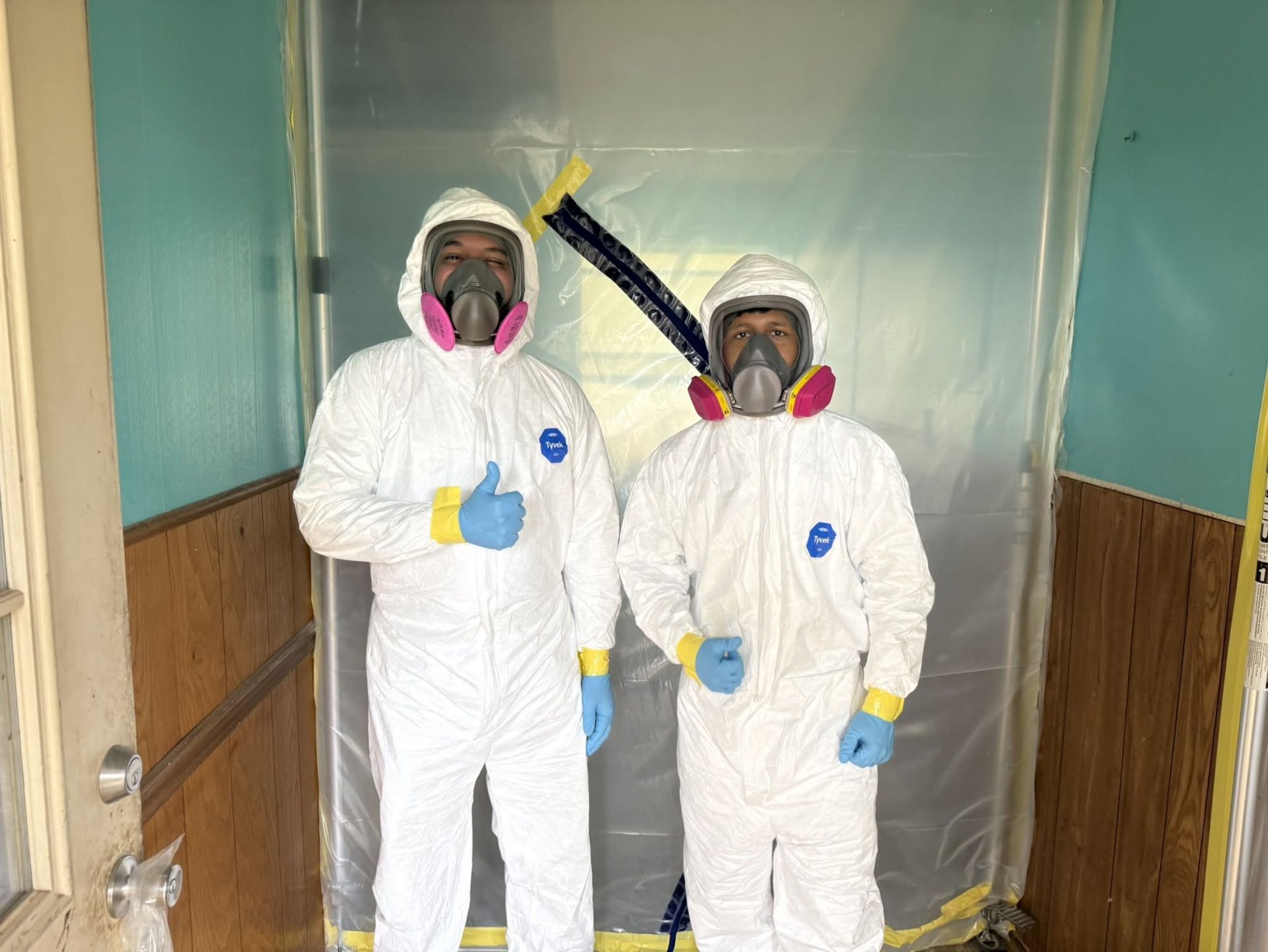 Two people in white hazmat suits and respirators stand in a plastic-lined hallway, one giving a thumbs-up.