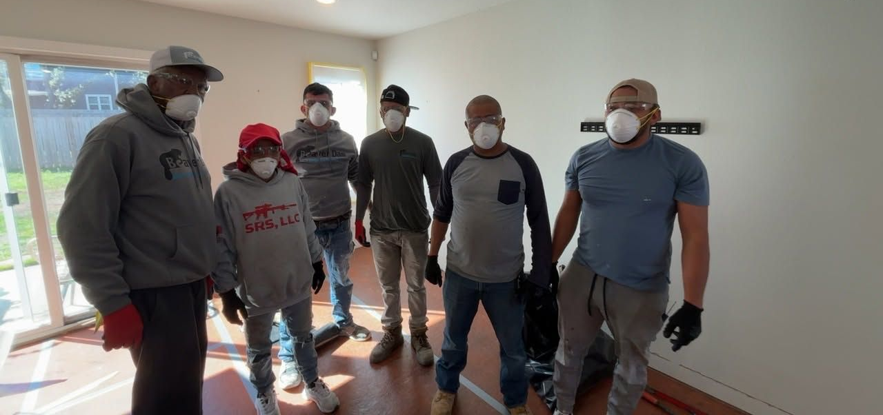 Five masked people stand indoors in a bright hallway, posing for the camera.