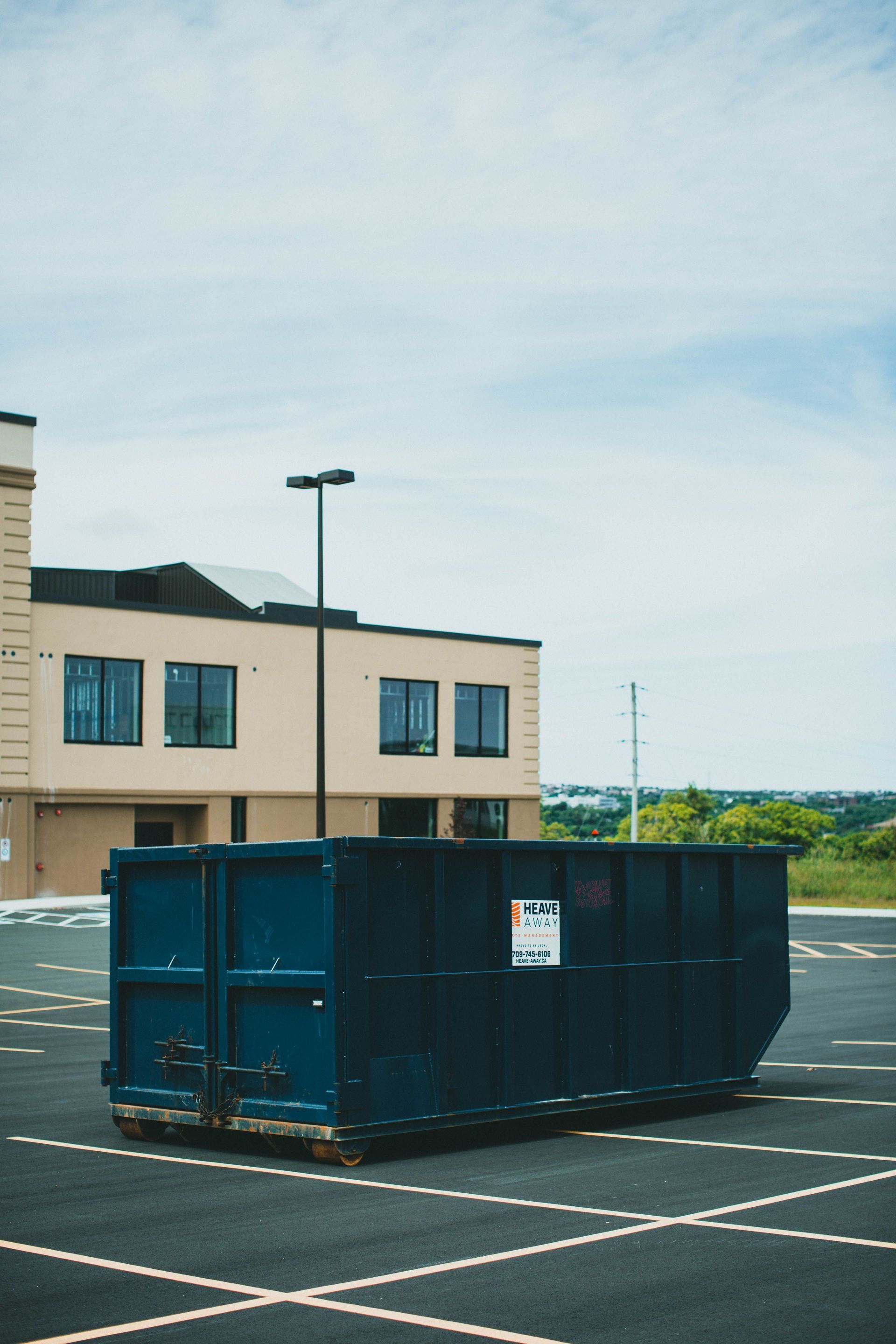 Blue dumpster in an empty parking lot beside a beige building under a cloudy sky