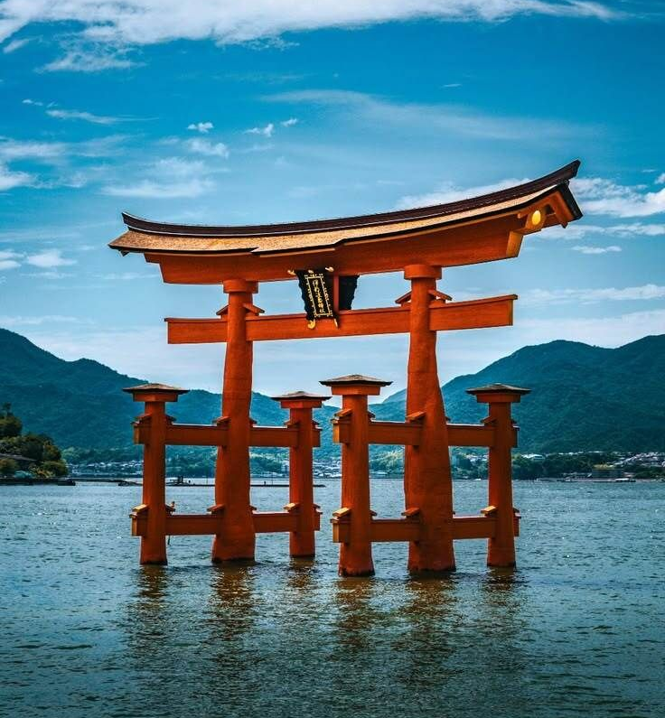Red torii gate standing in shallow water with mountains and cloudy sky in the background.