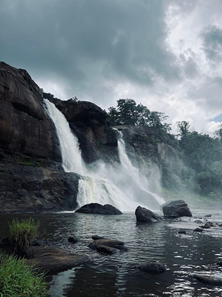Wide waterfall cascading over dark rocks into a misty river under cloudy skies