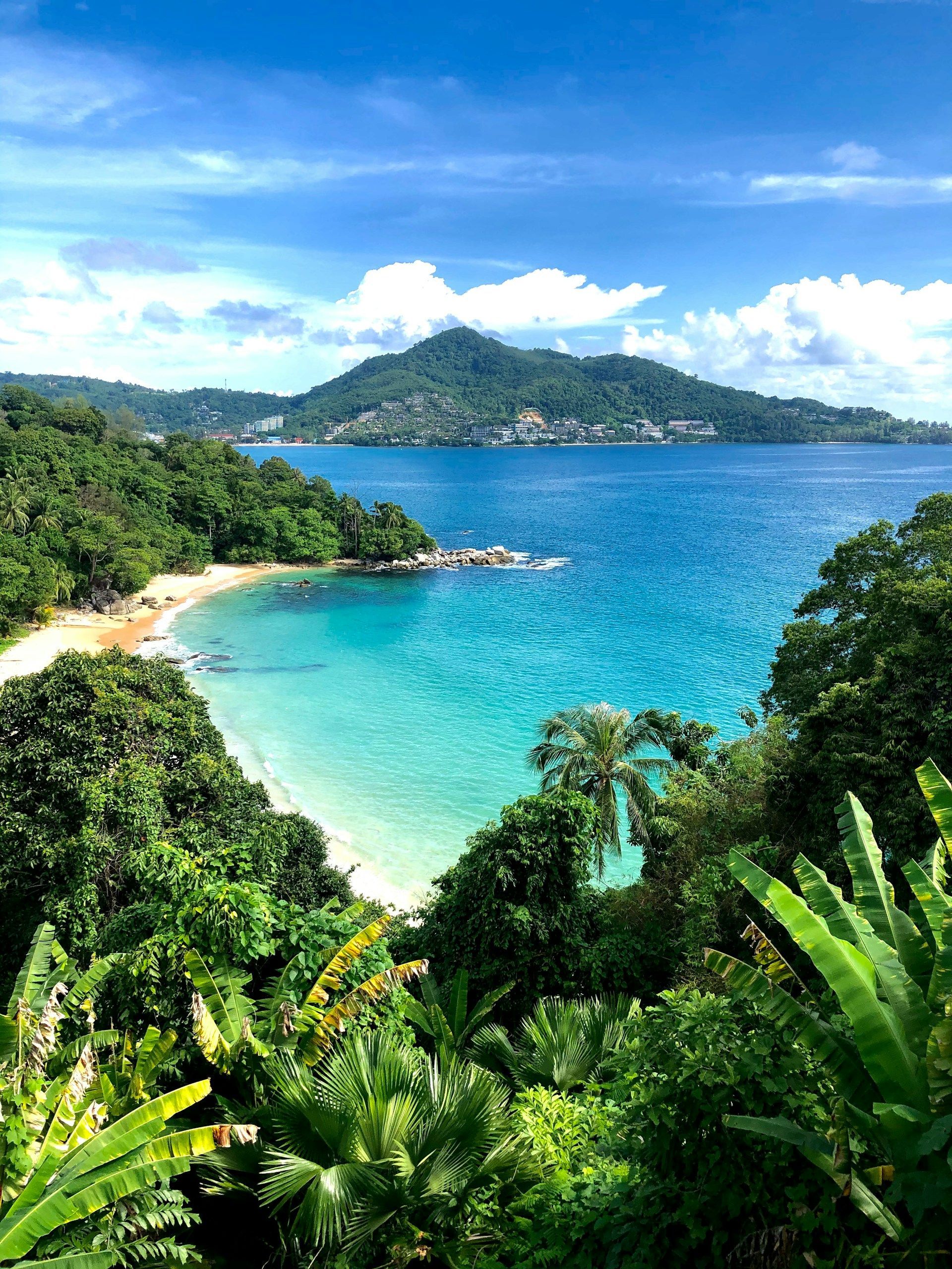 Turquoise bay surrounded by lush green foliage. A mountain and blue sky are in the background. Phuket,Thailand