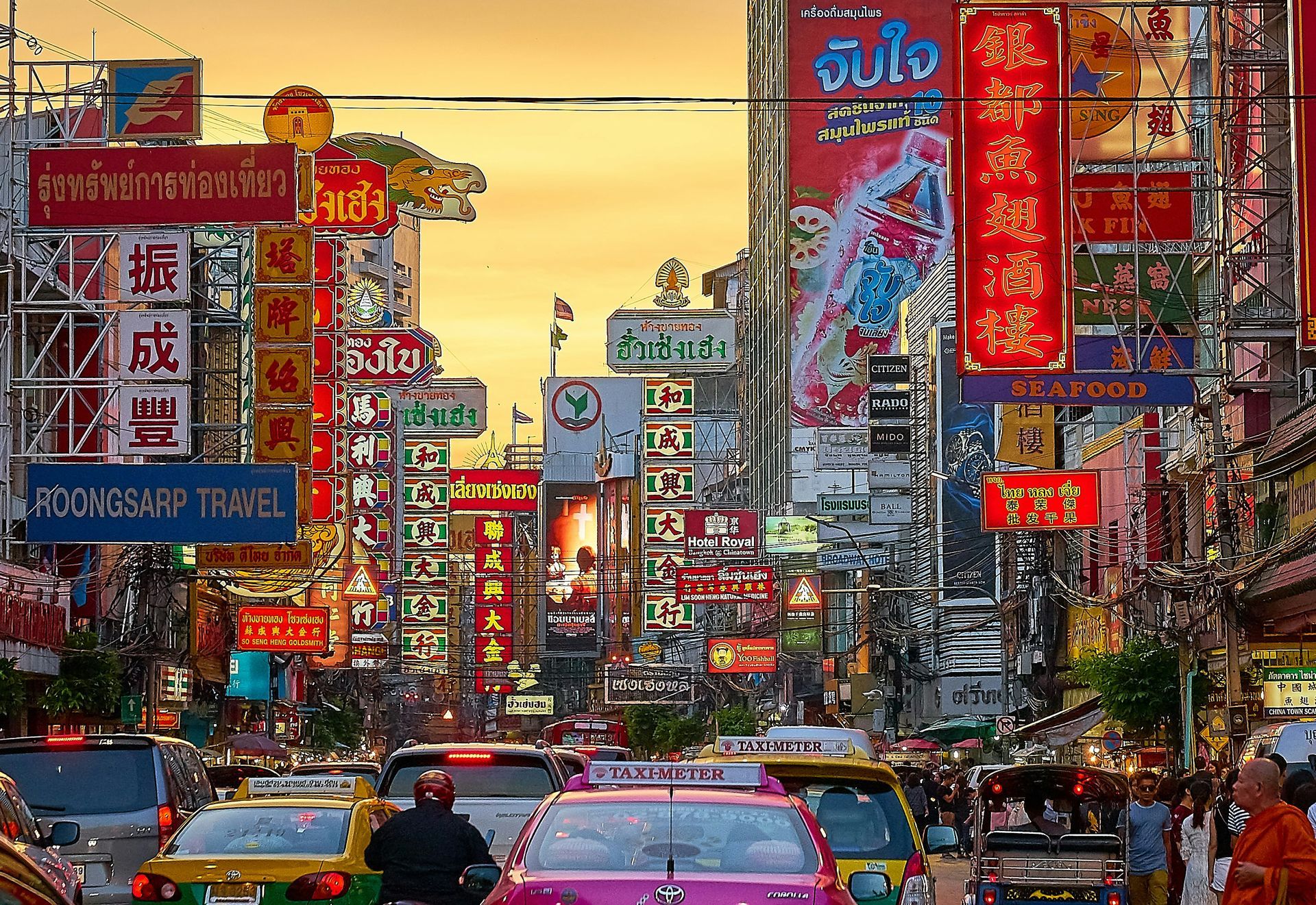 Busy city street lined with colorful billboards and neon signs, packed with traffic at sunset.