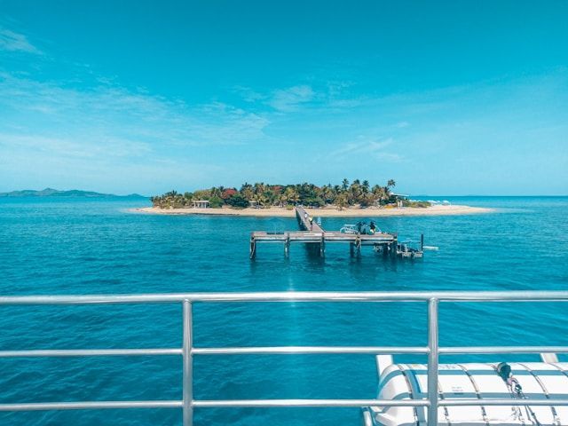 Island with lush greenery, blue water, and a pier, seen from a boat.