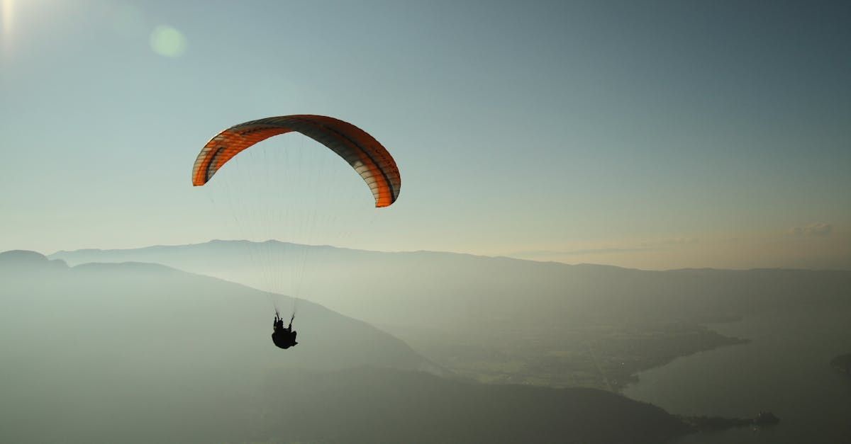 Paraglider soaring through a clear, blue sky over a hazy landscape of mountains and water.