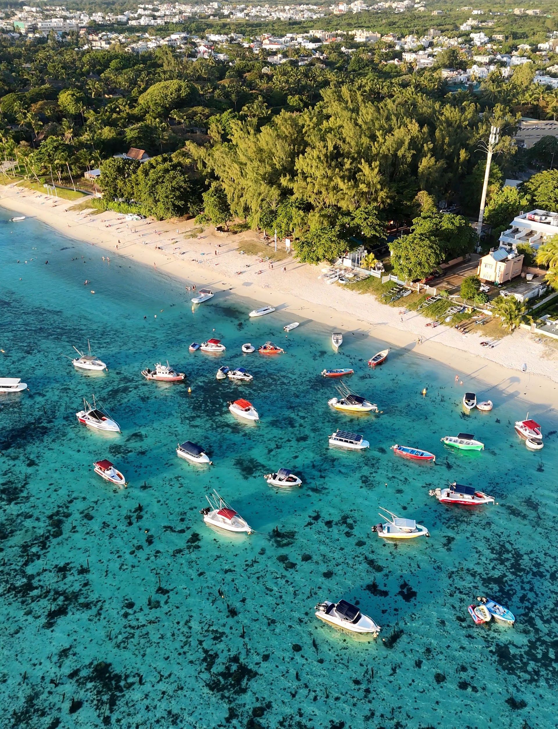 Aerial view of turquoise beach with many anchored boats and a tree-lined shore