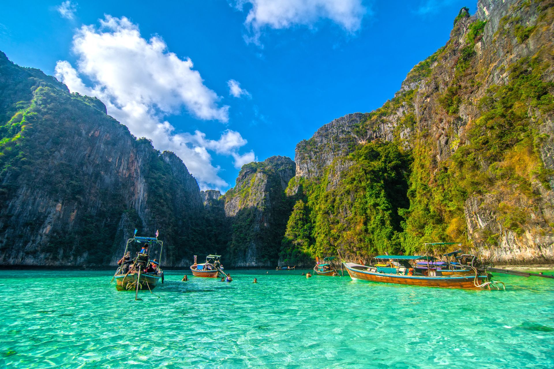 Turquoise lagoon with longtail boats beside steep green limestone cliffs under a bright blue sky