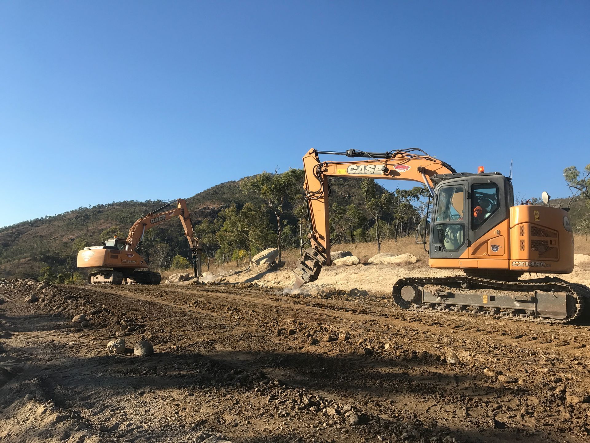 An Orange Excavator Is Working on A Dirt Road in The Woods — Watto's Earthmoving & Machinery Hire in Atherton, QLD