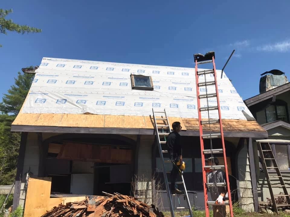 Workers installing white house wrap on a roof with a red ladder against a building under construction