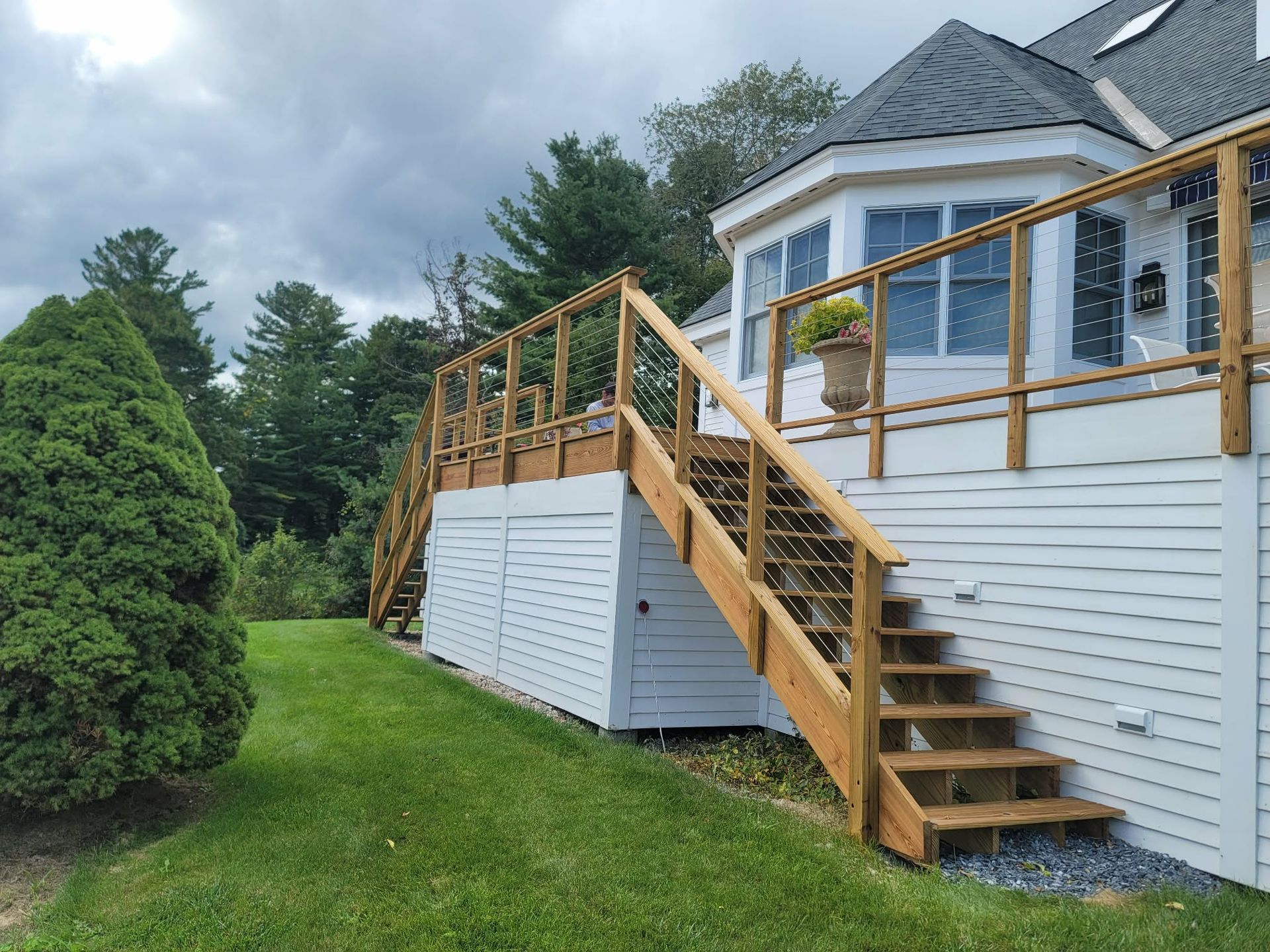 Wooden exterior stairs and deck on a white house beside a grassy yard and evergreen trees