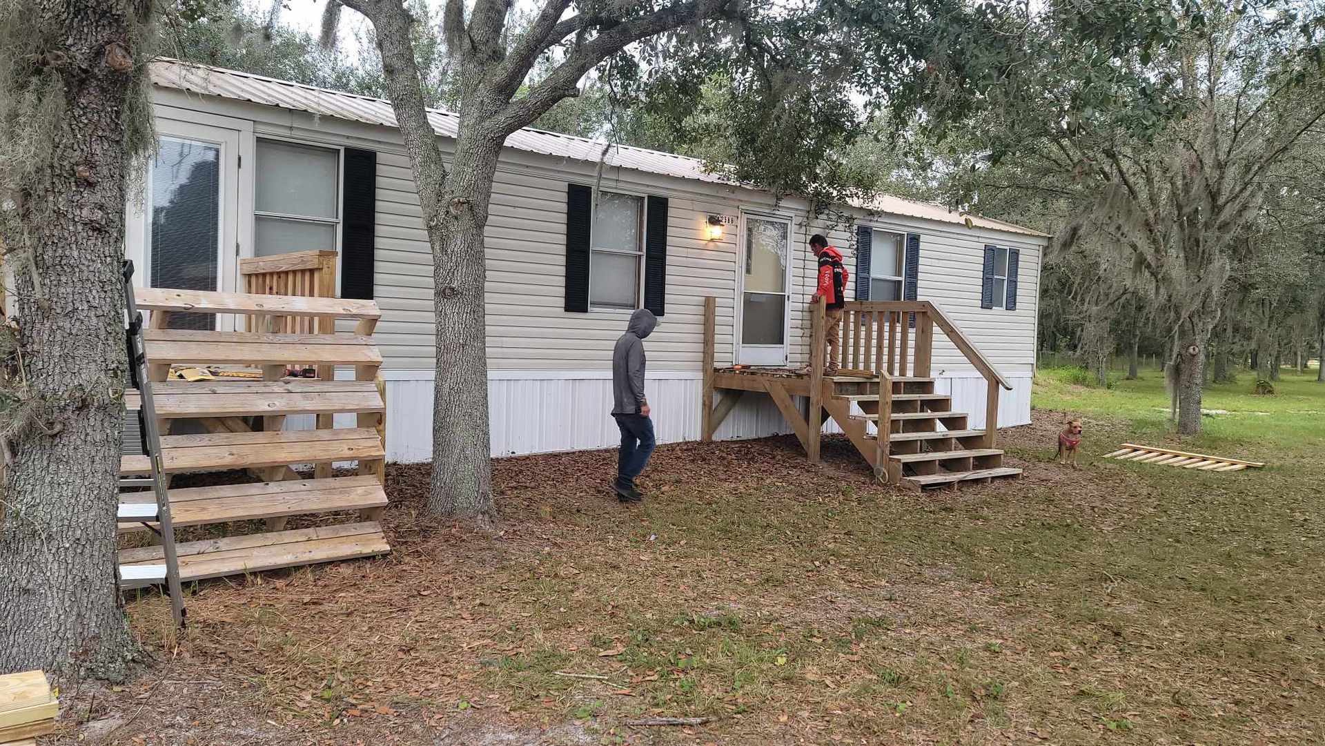 Single-wide trailer with wooden ramps and steps in a grassy yard, with a person standing outside.