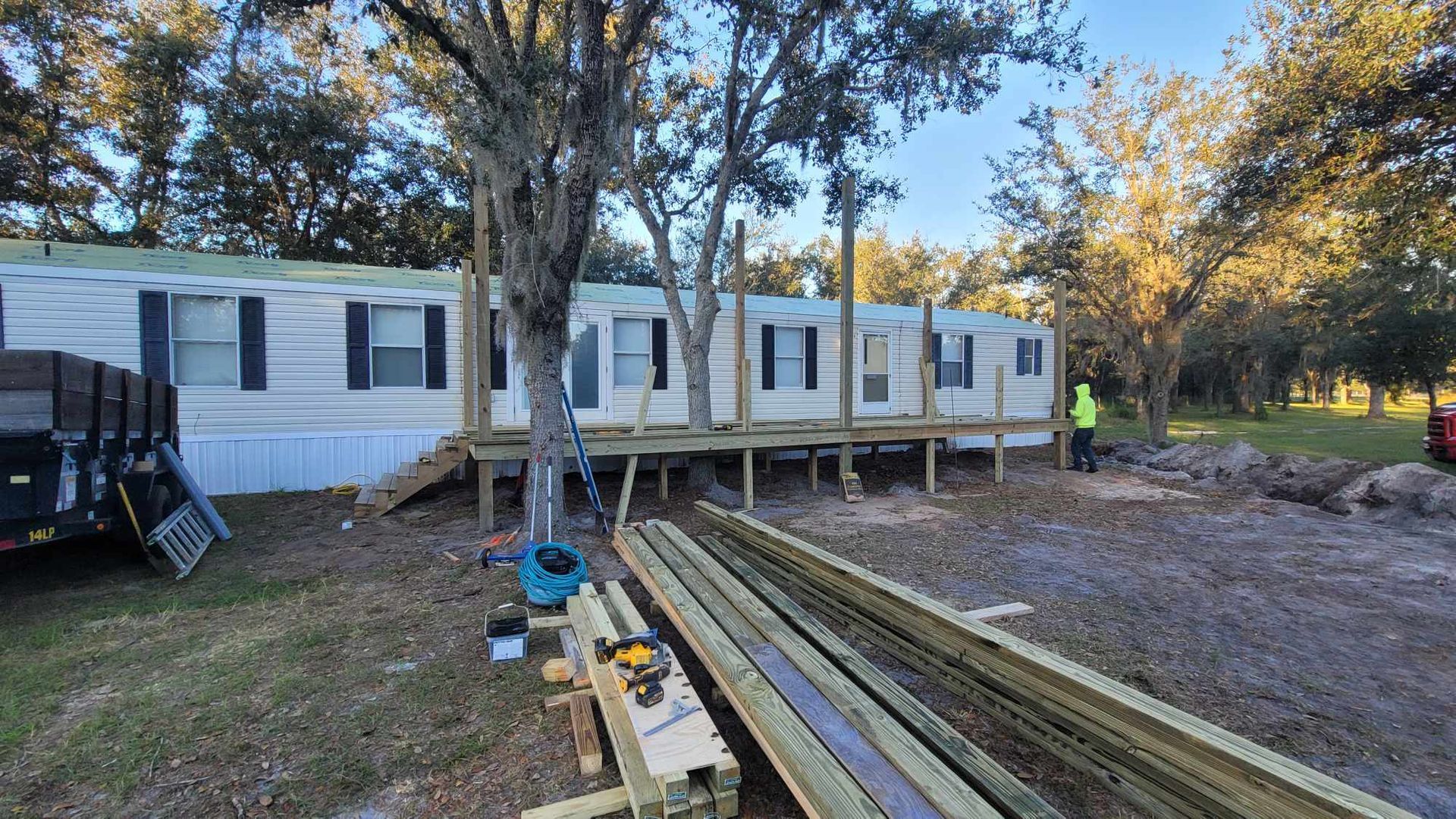 White manufactured home on raised supports in a wooded yard, with lumber stacked in the foreground.