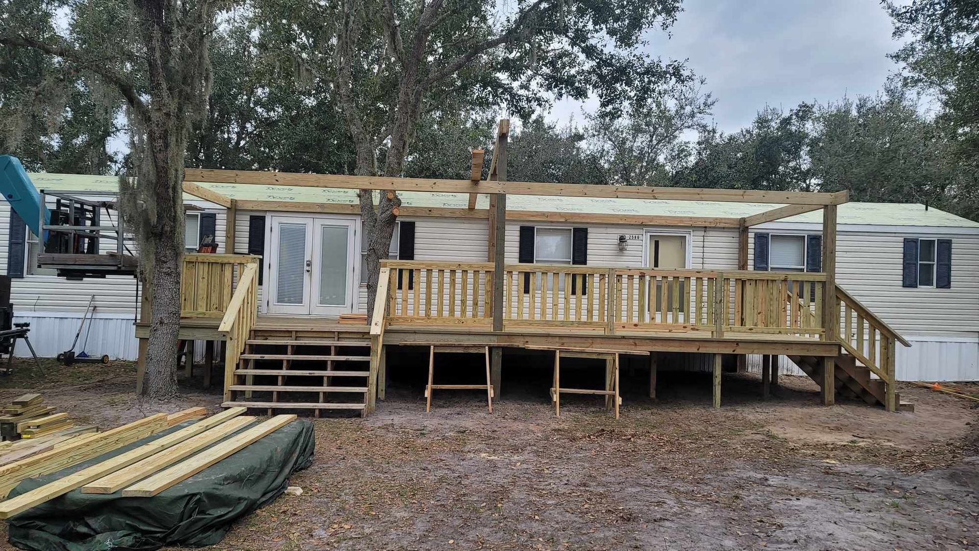 Weathered mobile home under construction on stilts, with a wooden porch and steps in a wooded lot.