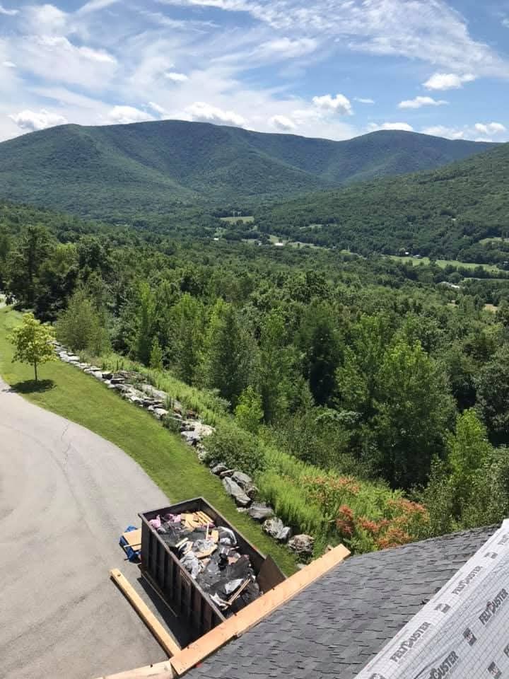 Mountain valley view with forested hills, blue sky, and a deck overlooking a road and parked vehicle