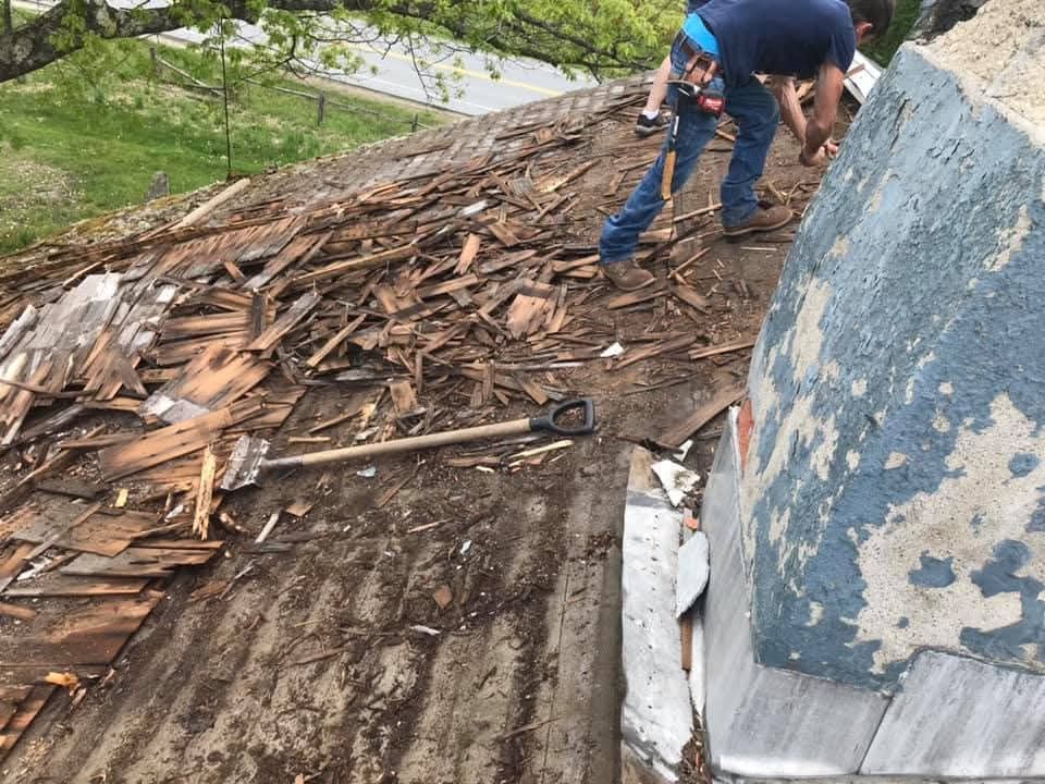 Workers repairing a damaged shingle roof beside a large blue wall.