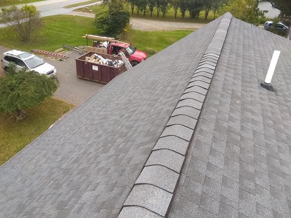 Roof shingles and ridge vent in foreground, with a dumpster and parked vehicles beside a grassy roadside.