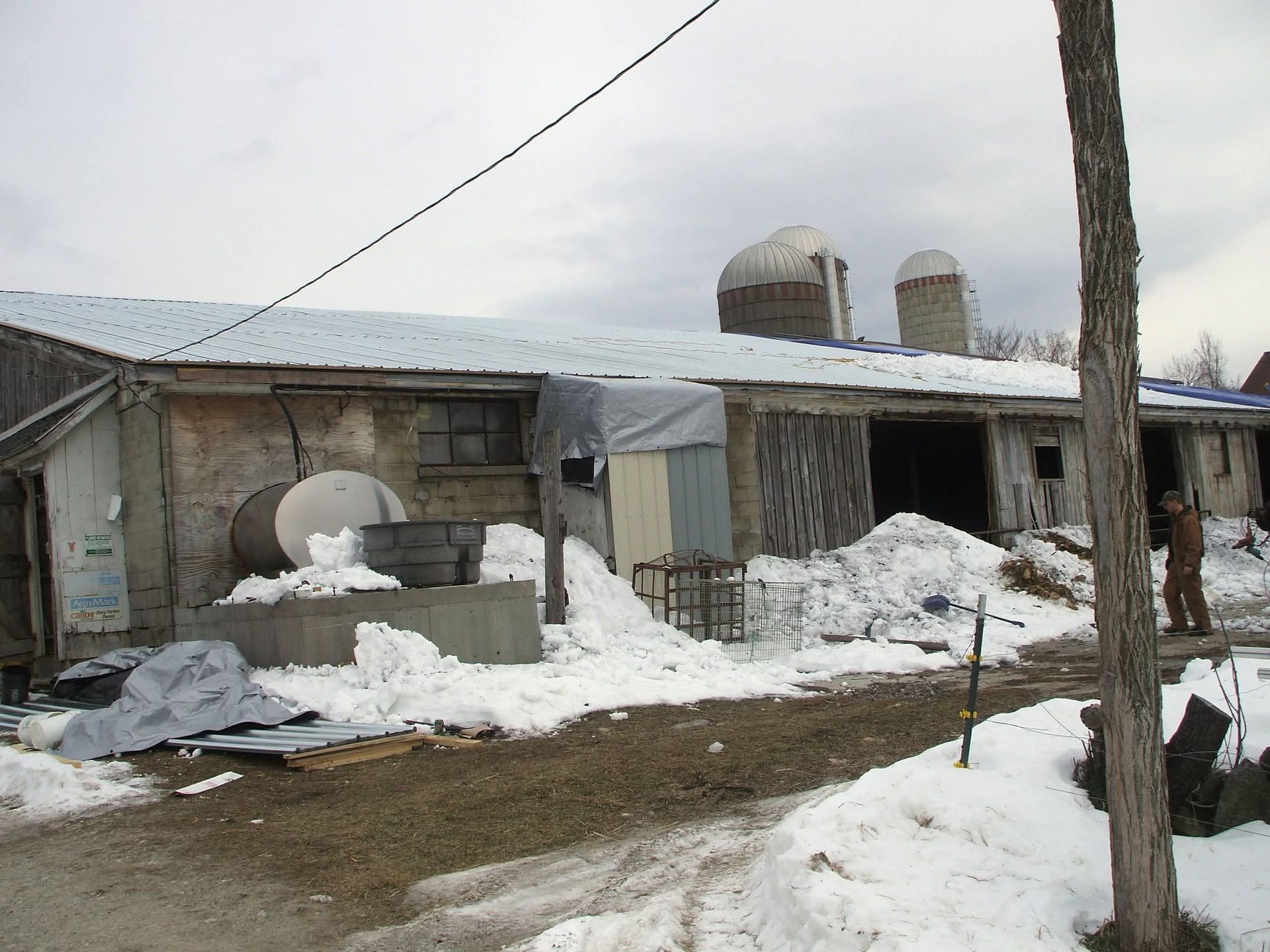 Snow-covered farmyard with a weathered barn, silos, and stacked materials under a cloudy sky