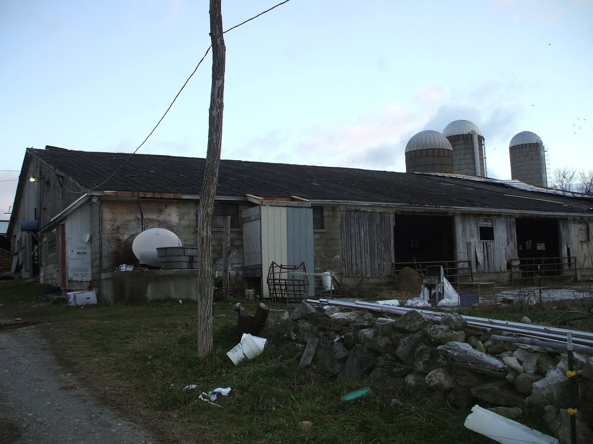 Dilapidated farmhouse with broken roof, silos, and scattered debris in a rural yard