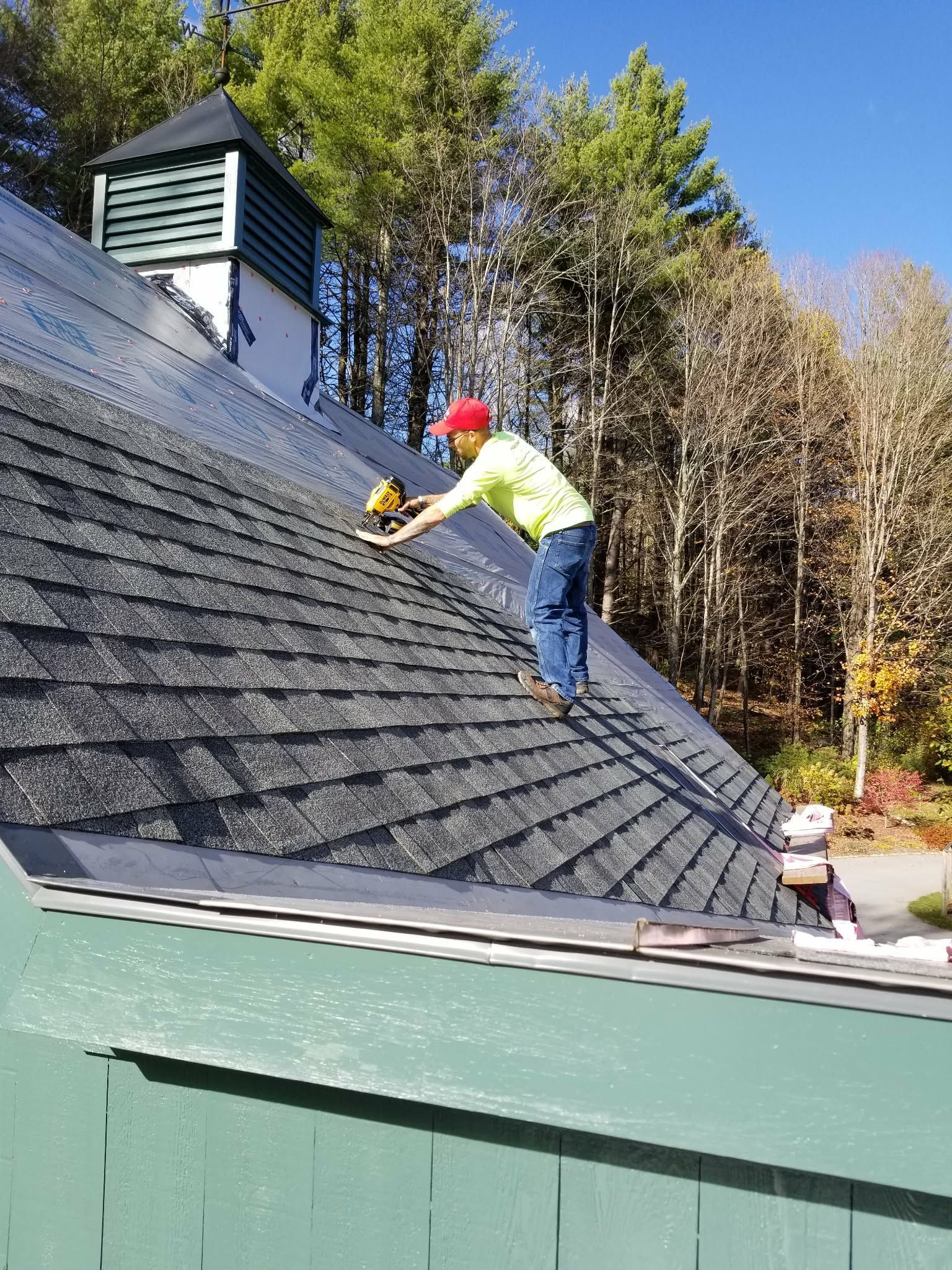 Worker on a steep shingled roof using a power tool, with trees and blue sky in the background.
