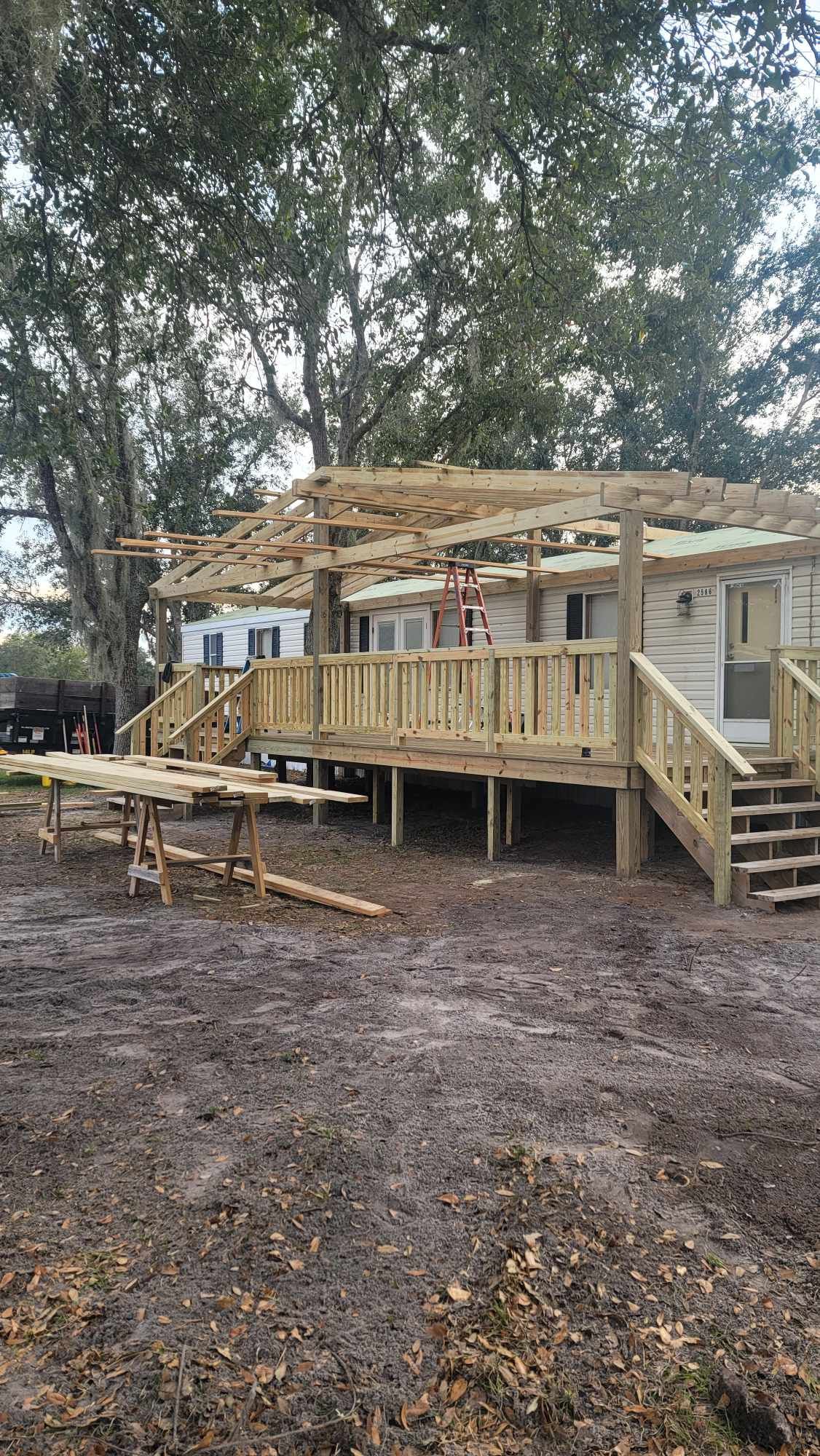 Wooden house under construction with deck, stairs, and roof framing in a wooded yard