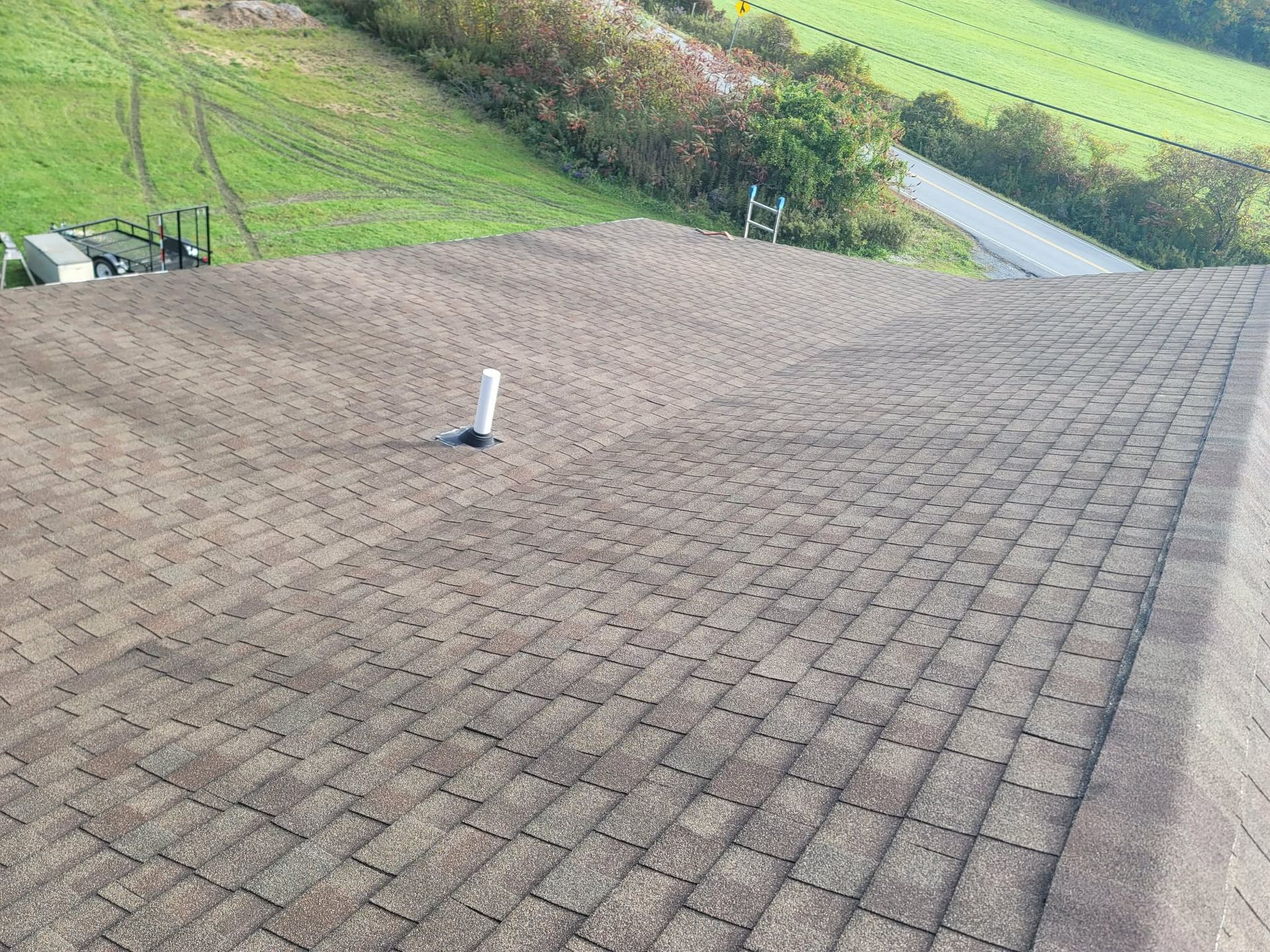 Roof shingles with a white vent pipe, overlooking green fields and a road beyond.