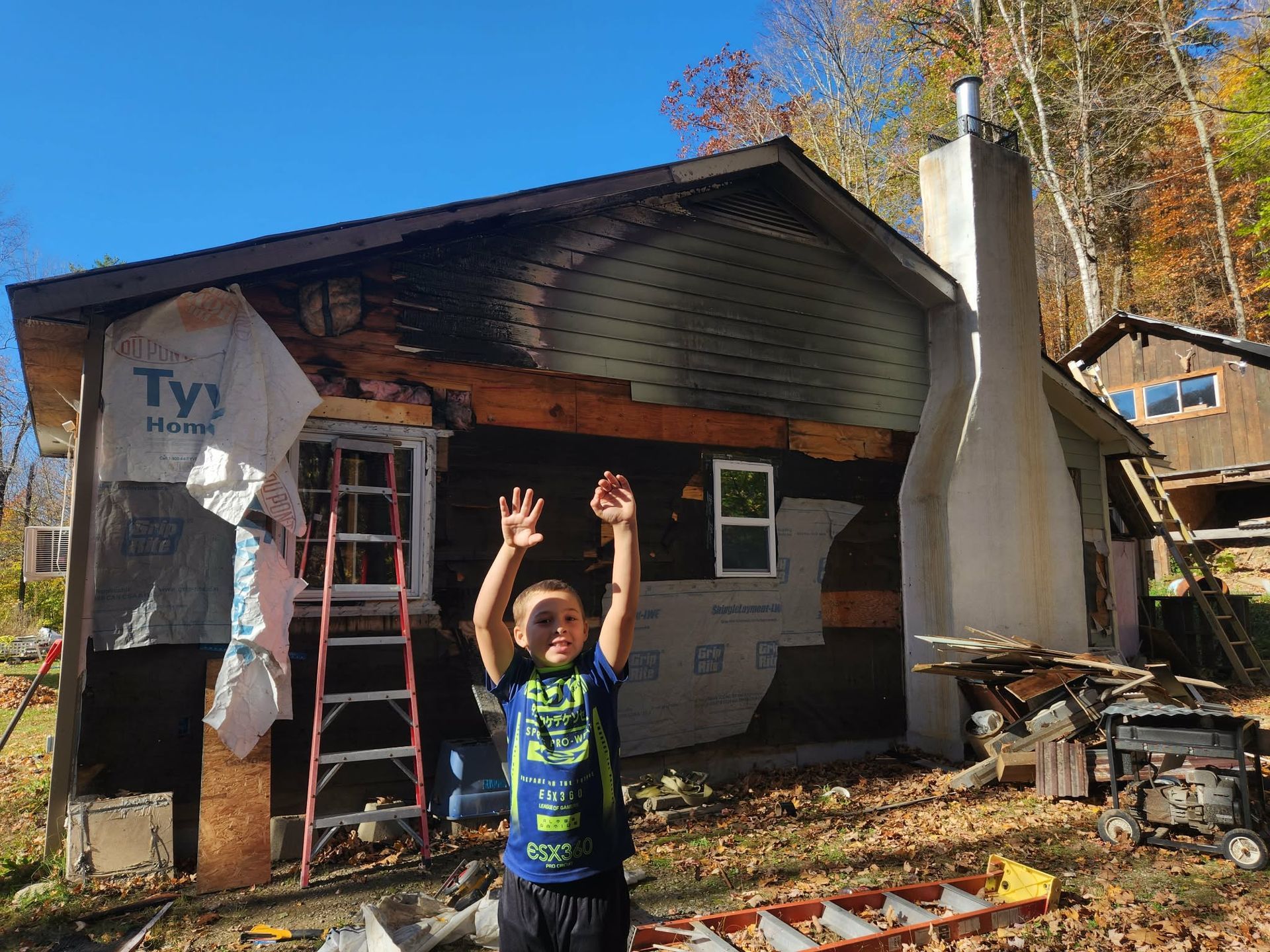 Child standing in front of a fire-damaged house with raised hands and a ladder nearby