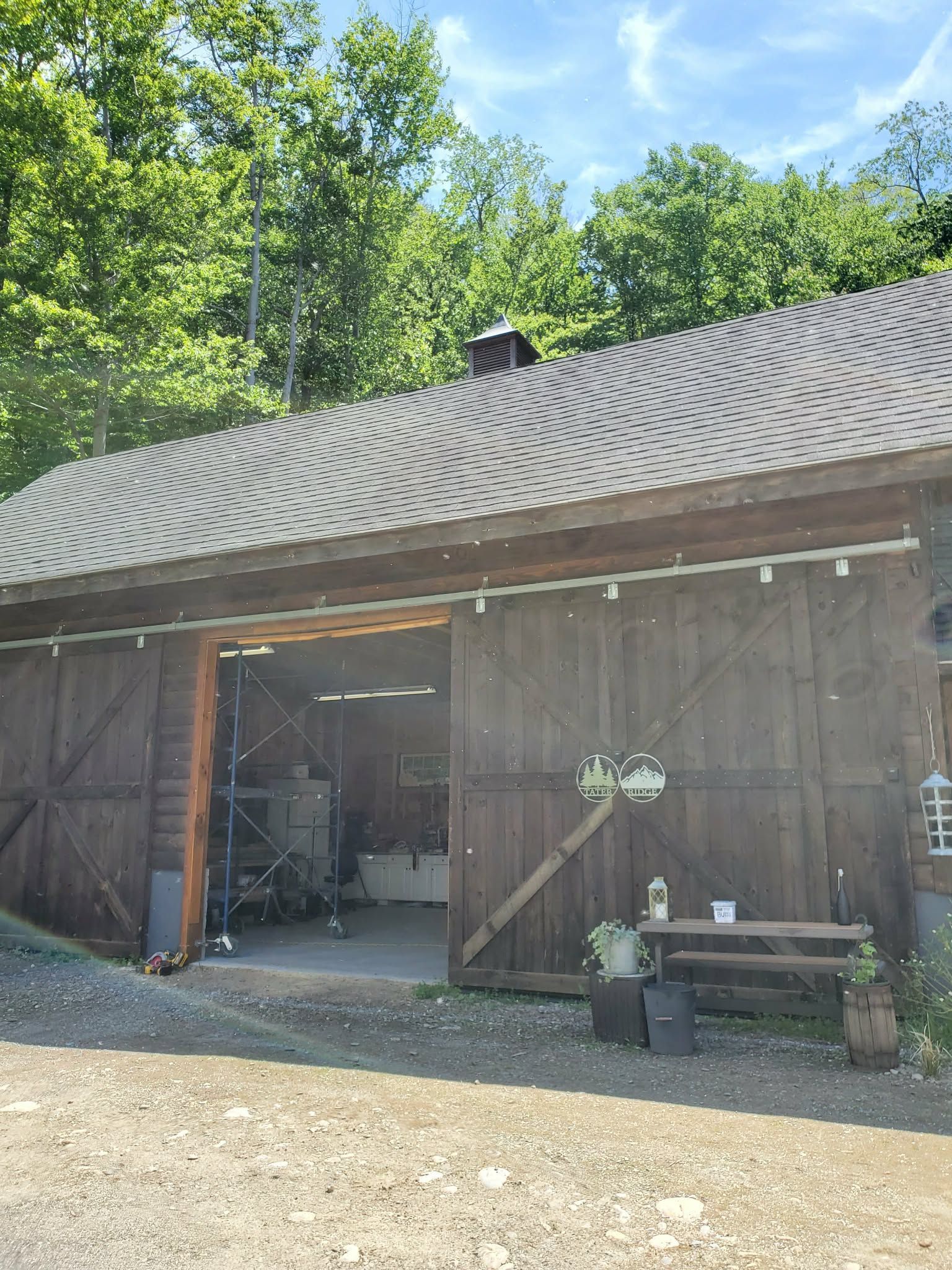 Rustic wooden barn with open doors, patterned roof, and potted plants in front, surrounded by trees.