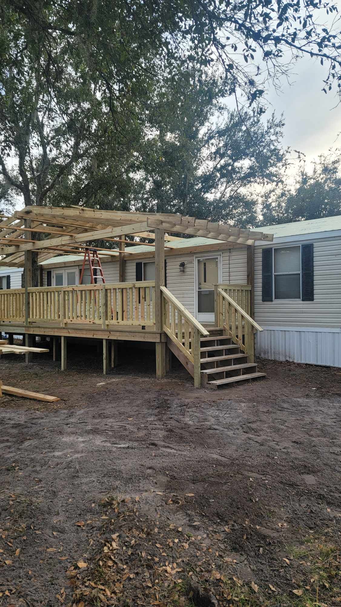 Small wooden porch with steps attached to a beige mobile home in a wooded yard
