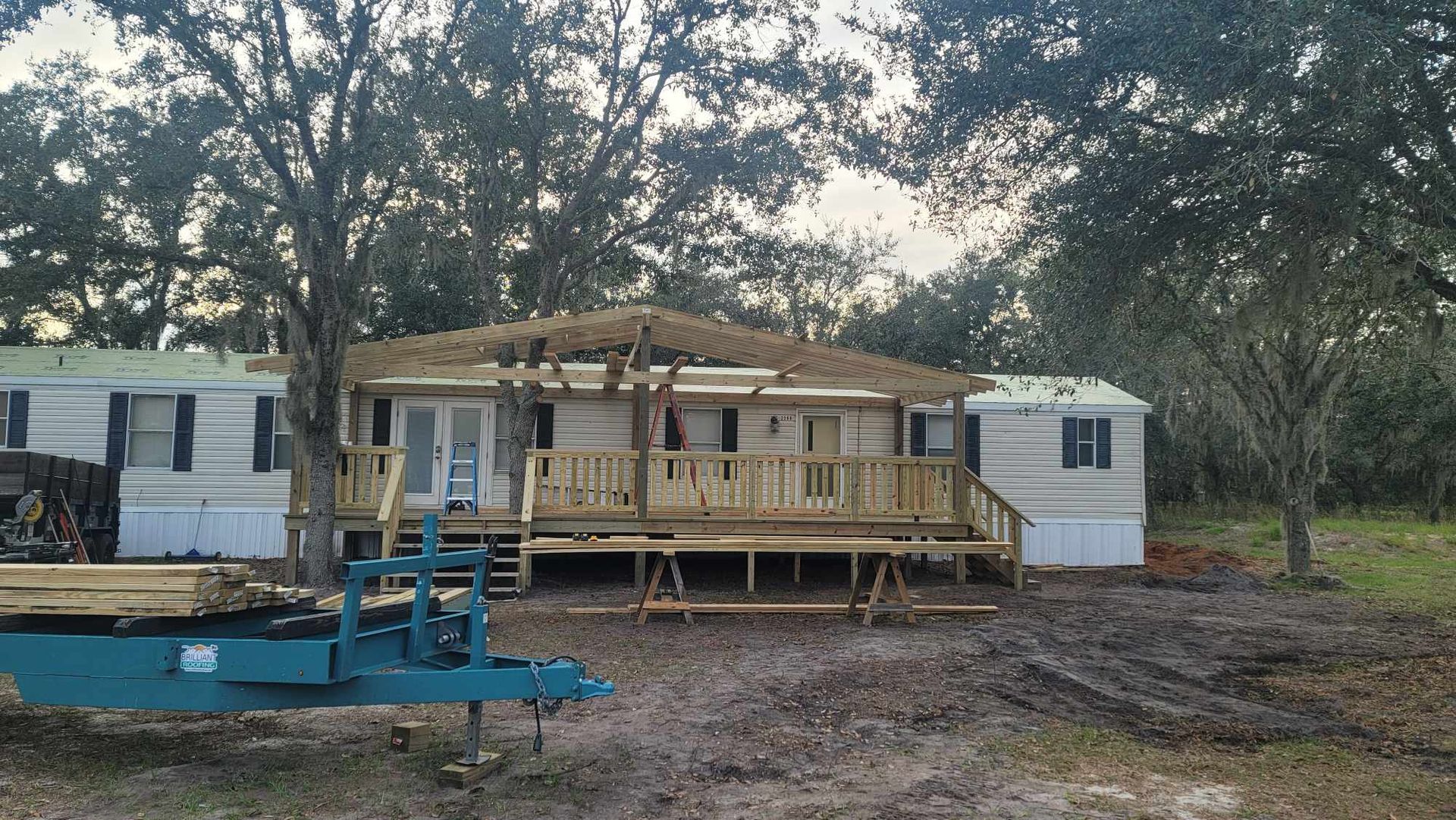 Partially built wooden house with porch, flanked by two white mobile trailers in a wooded yard