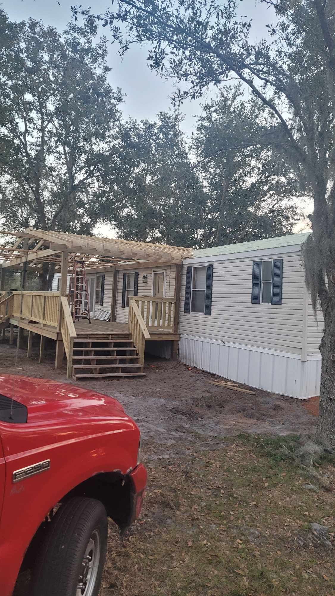 White mobile home with a wooden porch and steps beside a red truck under tall trees