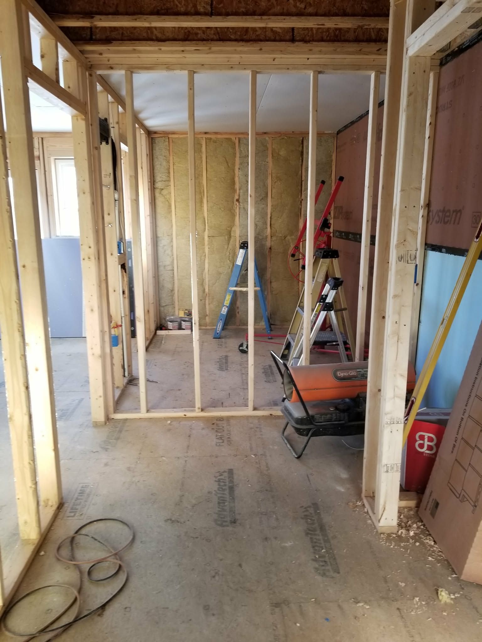 Interior under construction with framed walls, exposed wood, tools, and a wheelbarrow on a concrete floor.