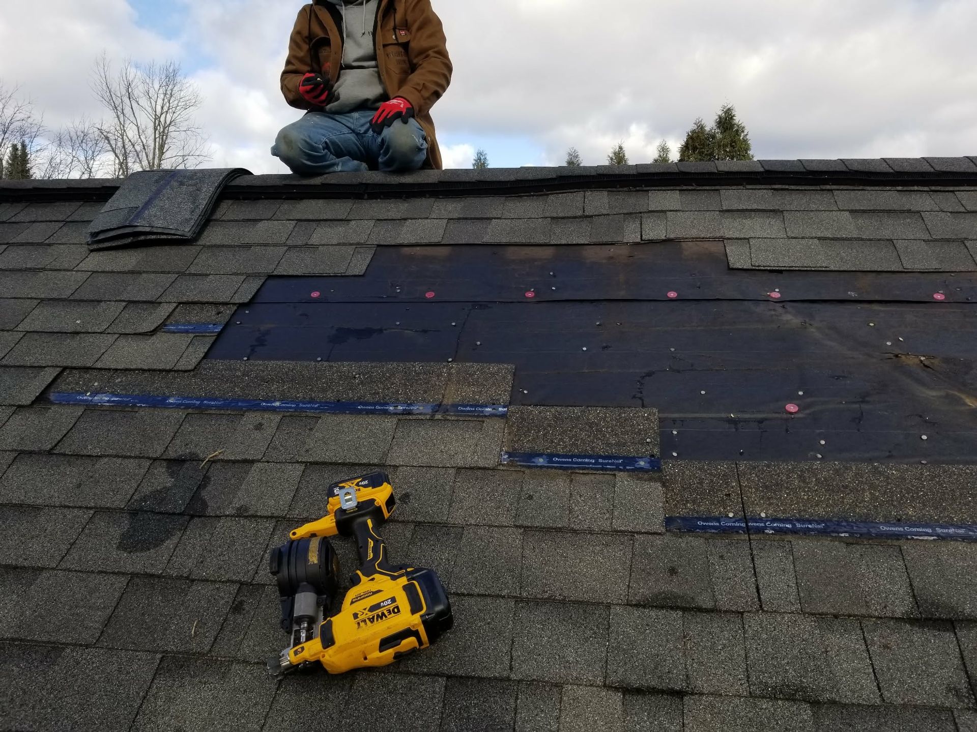 Worker on a shingled roof examining a repair area with a yellow power drill nearby.
