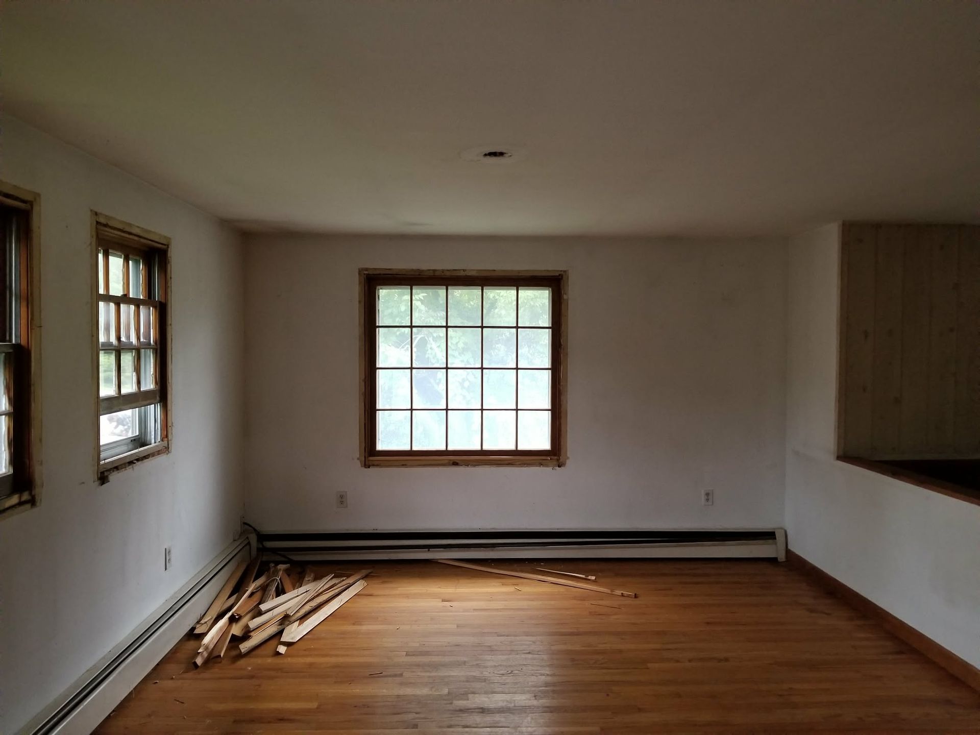 Empty room with wood floors, white walls, windows, and stacked lumber along the left wall.