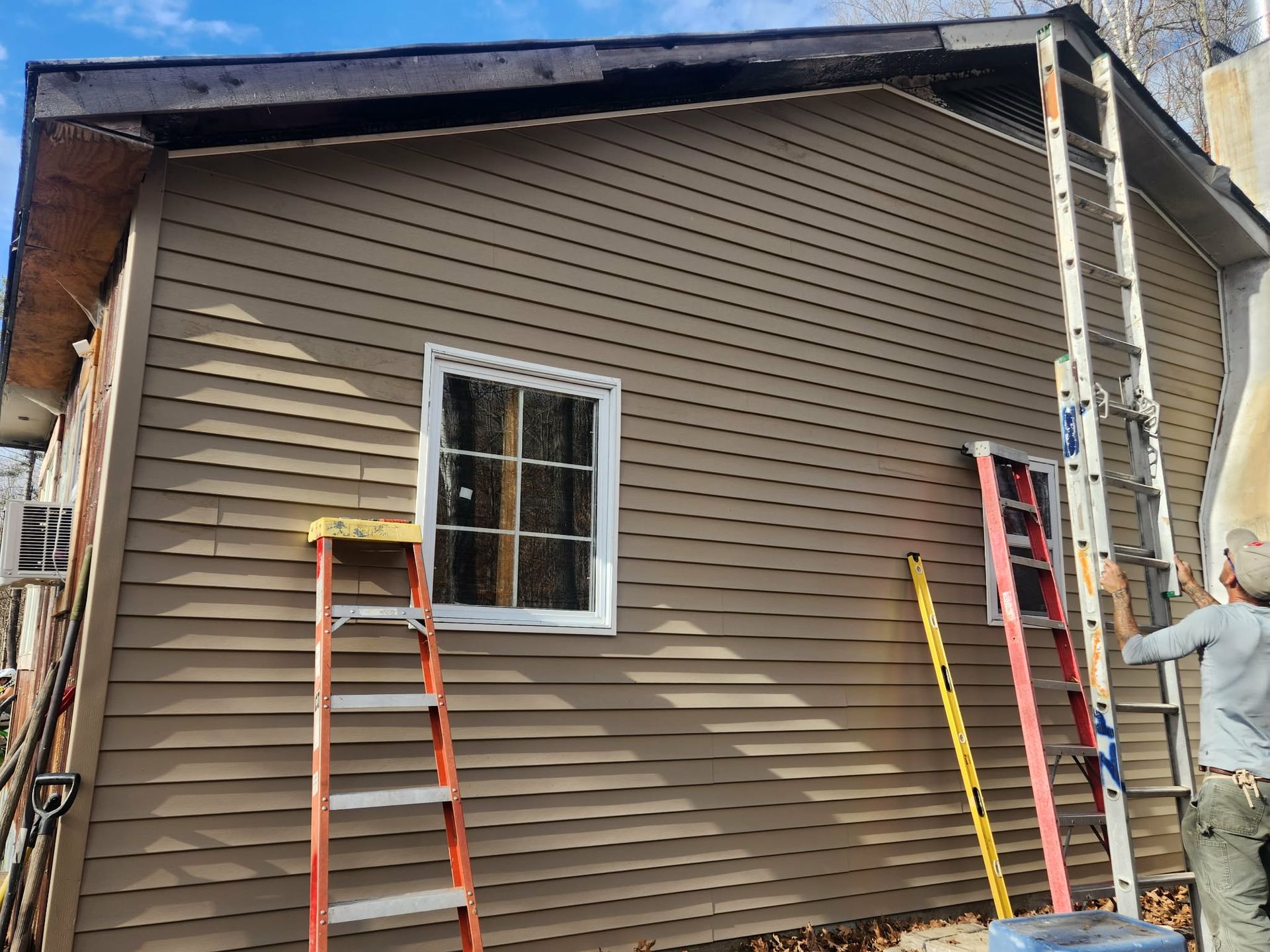 House exterior with tan siding, a window, ladders, and a worker painting trim.