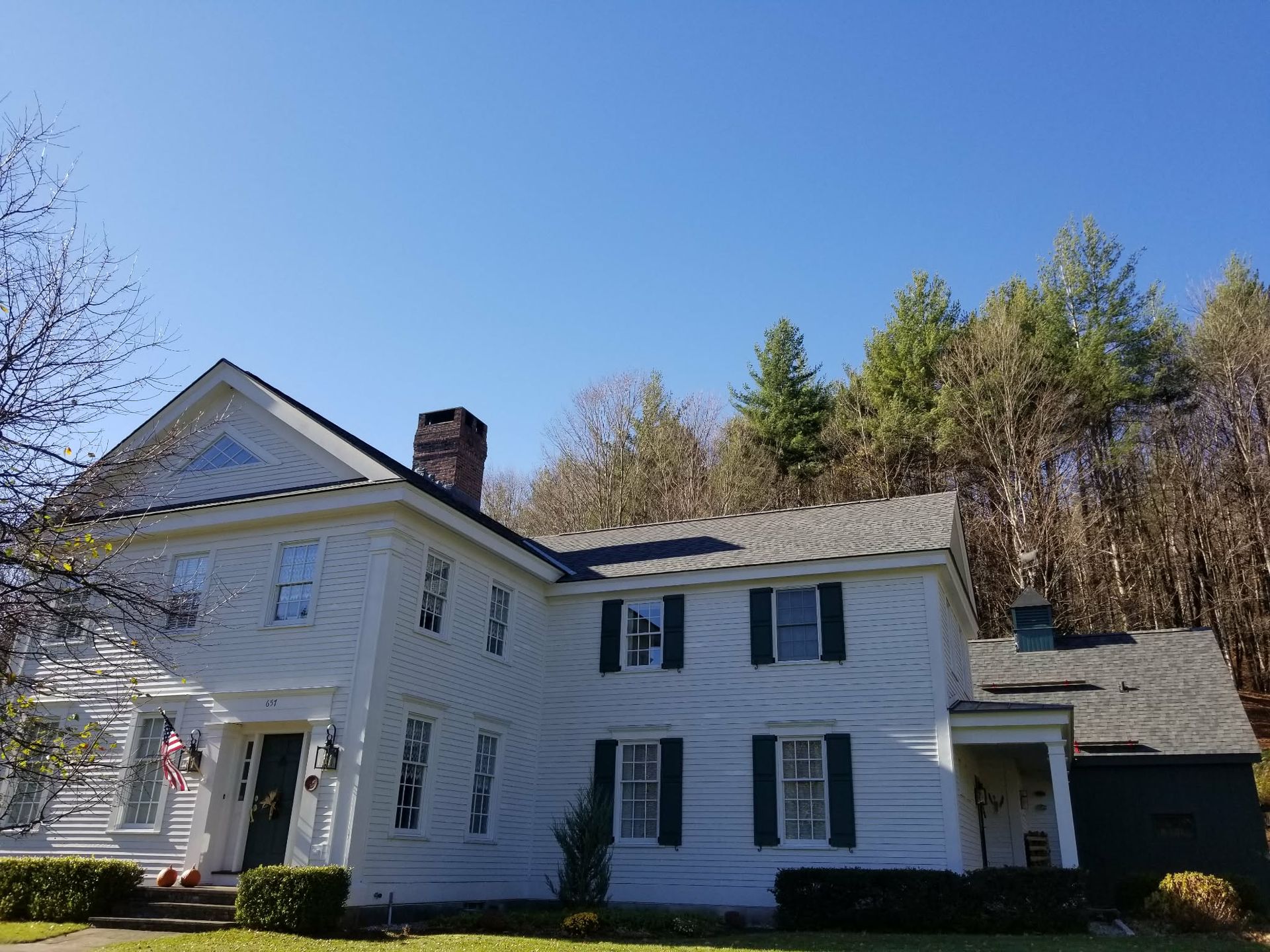 White farmhouse with blue shutters and a porch, set against trees under a clear blue sky