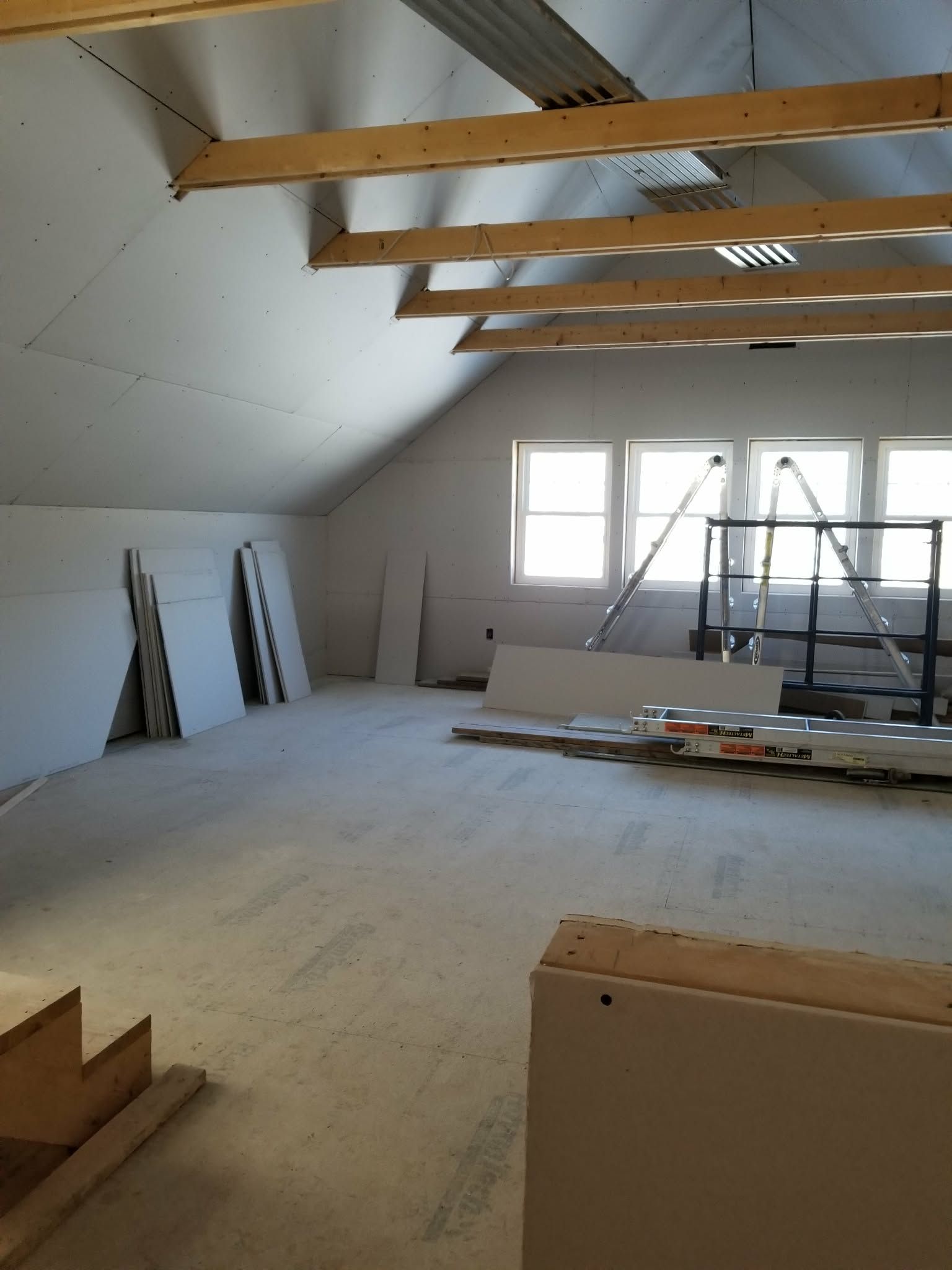 Attic room under renovation with exposed beams, white walls, windows, and construction materials on the floor