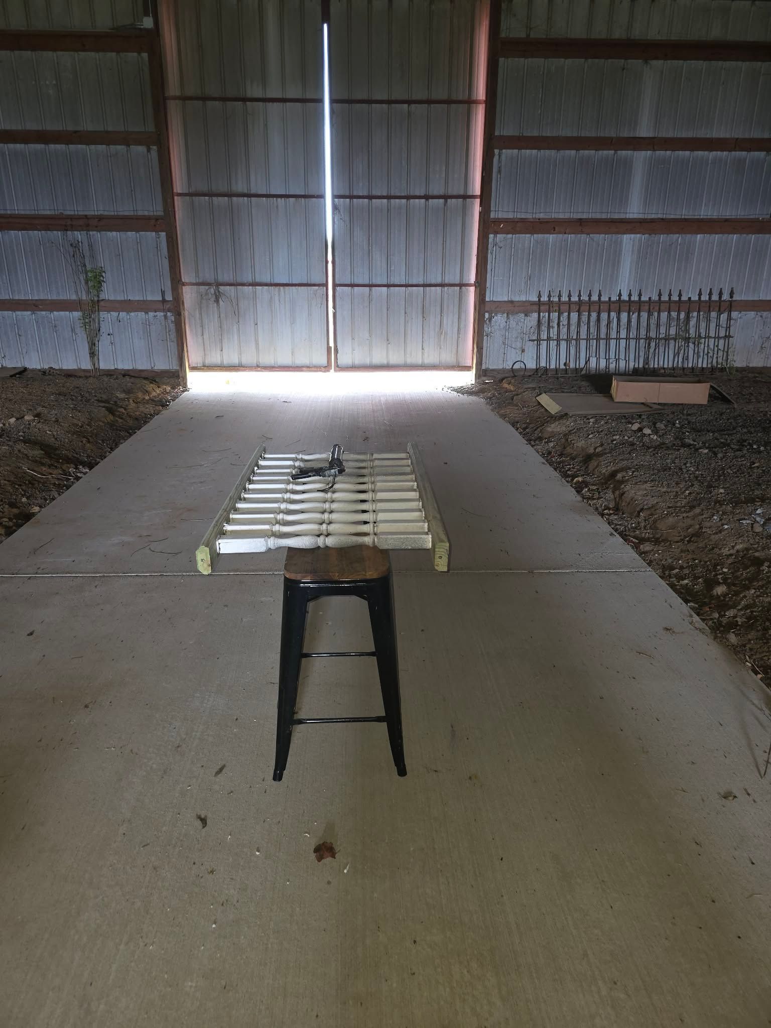 Metal stool centered on a concrete floor in a dim barn, with bright light streaming through open doors.